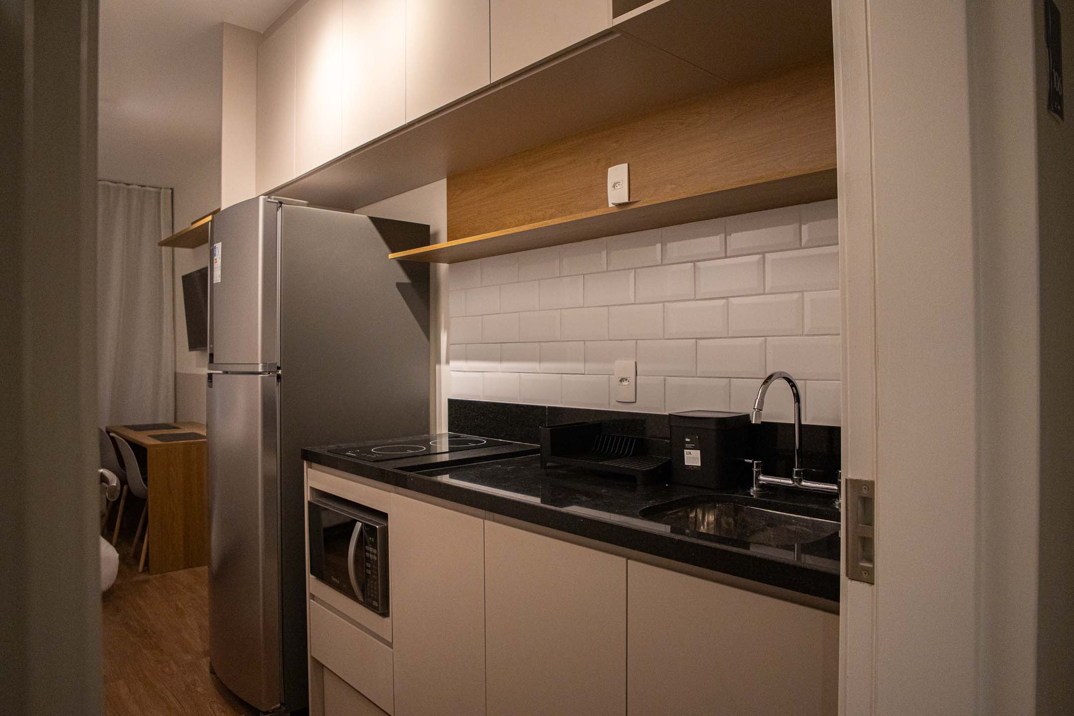 Cozinha com bancada de granito preto, cooktop embutido, cuba de inox, revestimento metrô e geladeira inox
