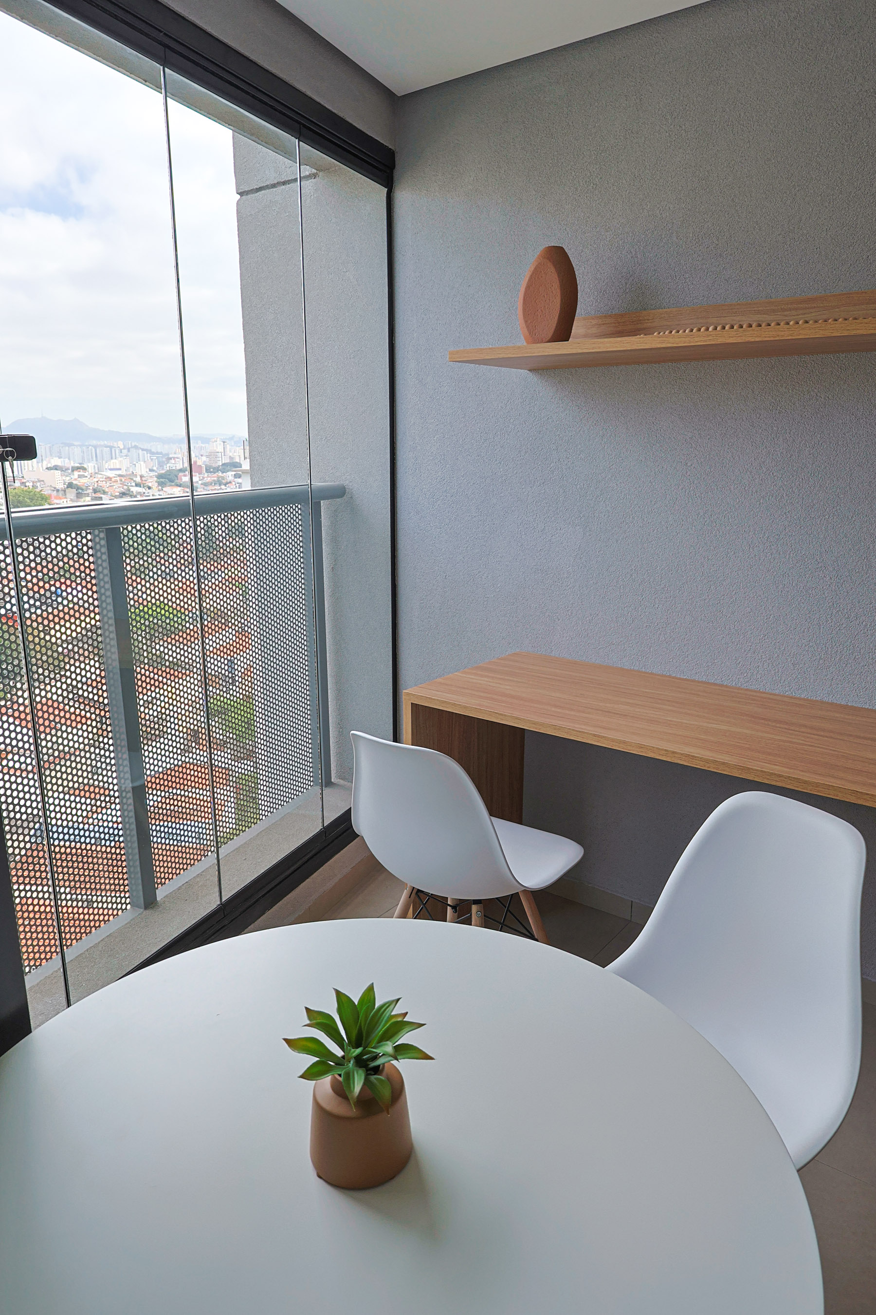 Varanda envidraçada com mesa redonda branca, cadeiras, bancada de madeira e vista da cidade