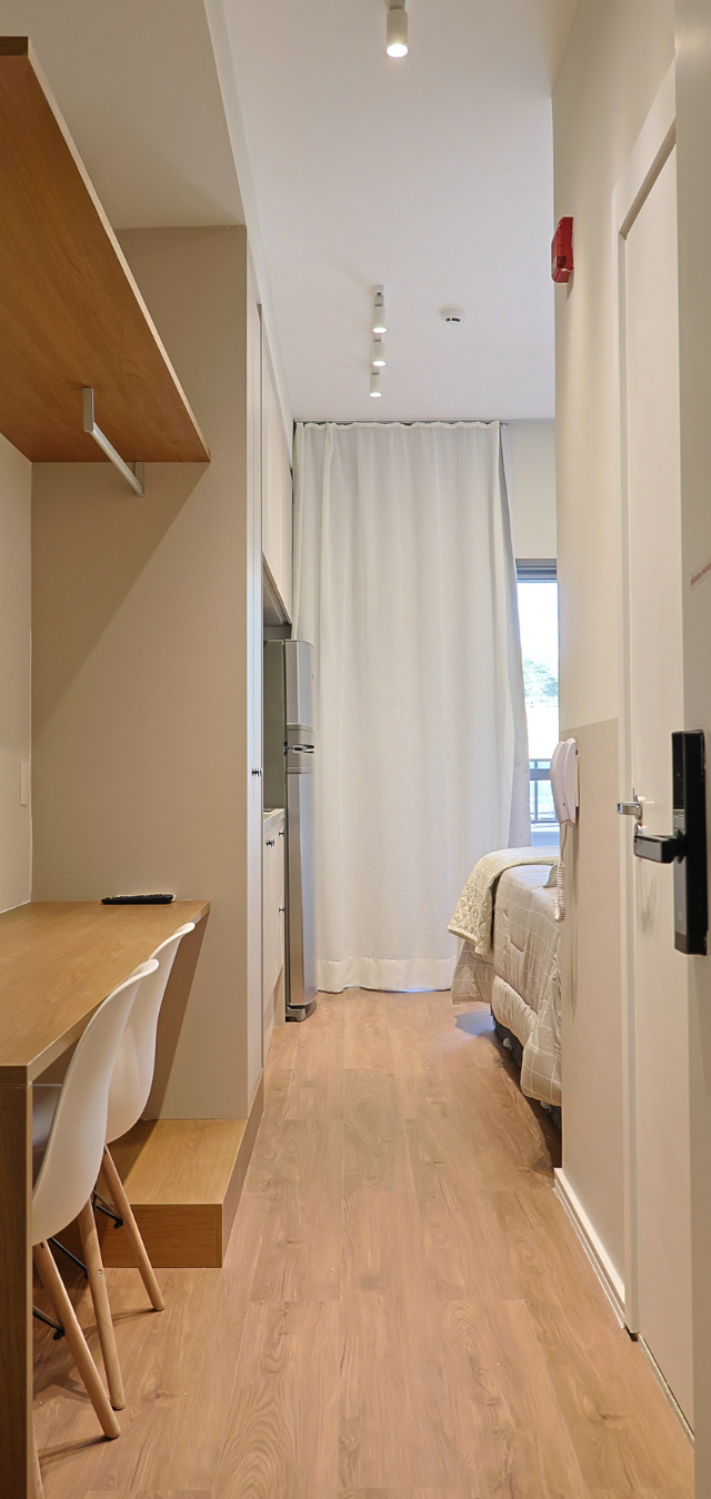 Obra pronta: Corredor do studio com bancada de madeira, cadeiras brancas, porta com fechadura digital e cama ao fundo