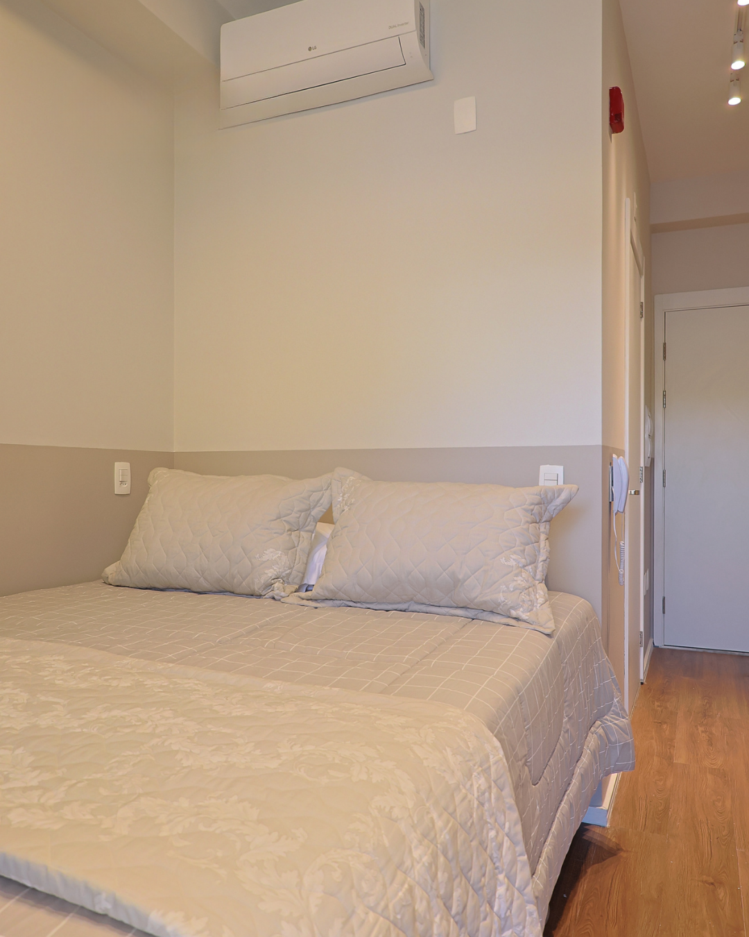 Obra pronta: Cama de casal com roupa de cama clara, meia-parede bege e ar-condicionado acima