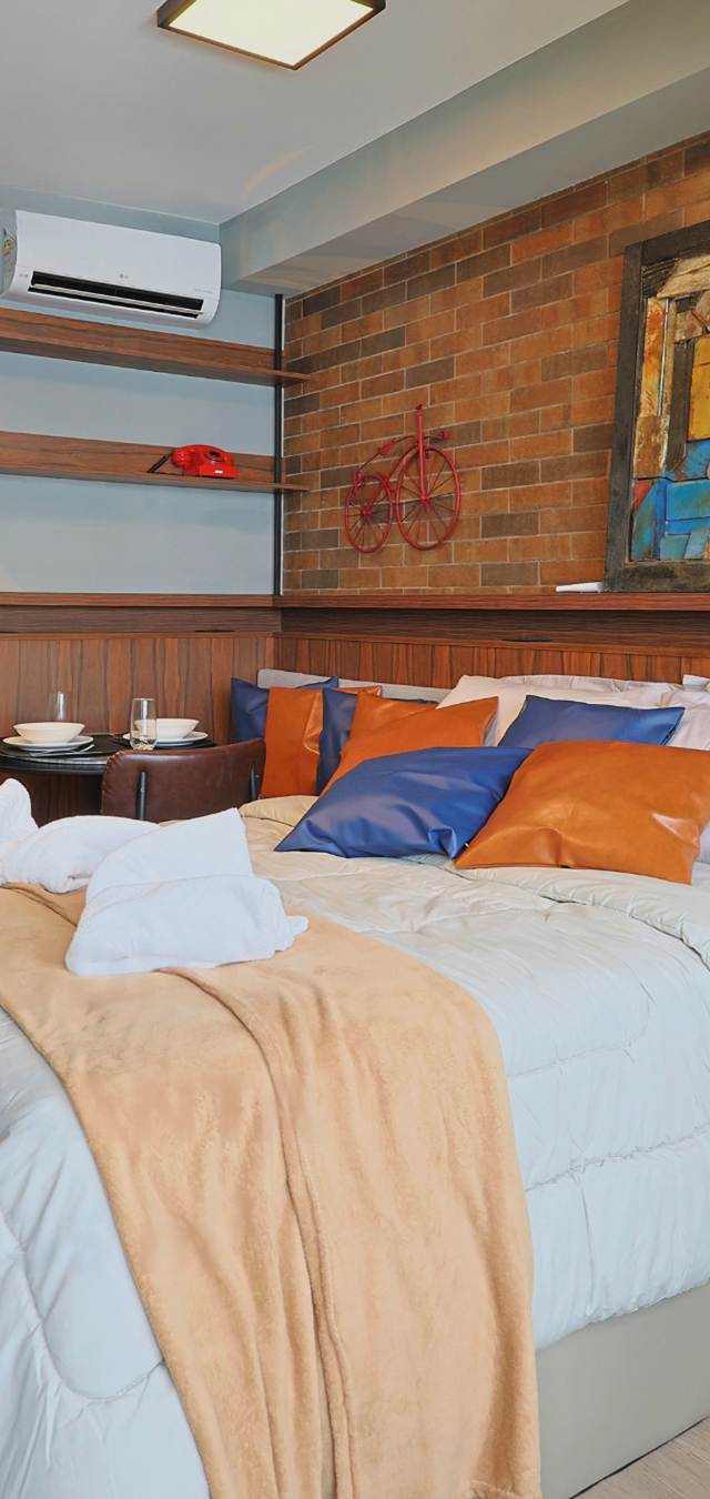 Obra pronta: Cama de casal com almofadas laranja e azuis diante de parede de tijolinho com quadro e bicicleta decorativa