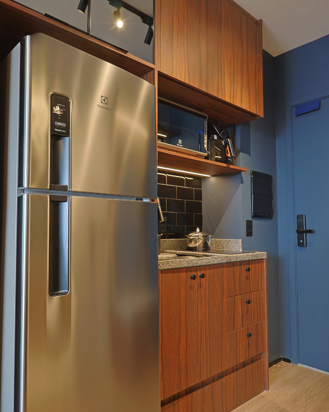 Obra pronta: Cozinha com geladeira inox, armários de madeira escura, metrô preto e parede azul com porta de entrada