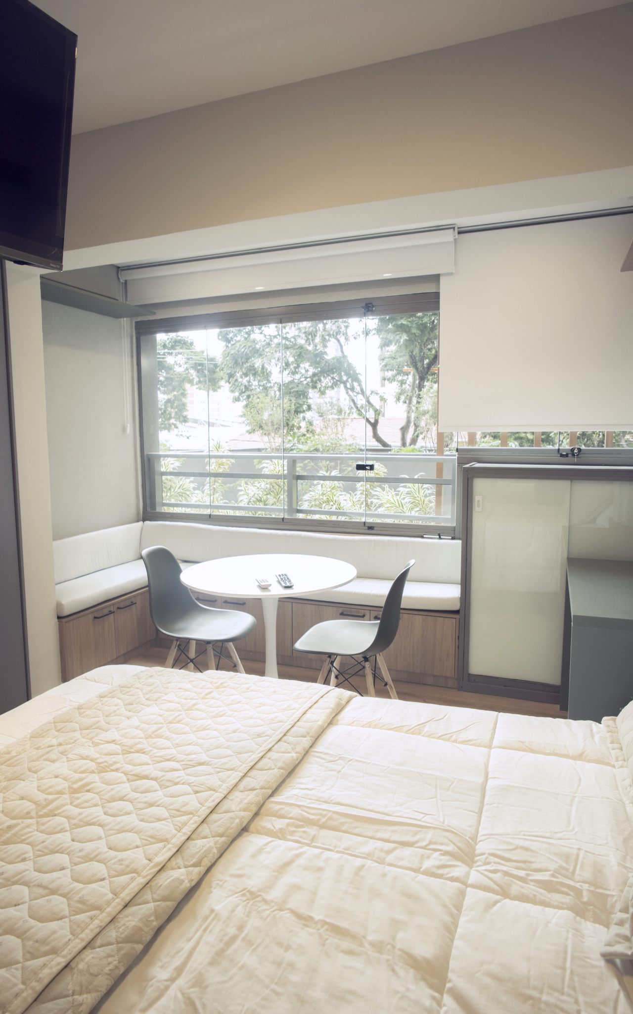 Foto do Latitude Campo Belo com cama em primeiro plano, mesa redonda diante da janela e banco com assento claro ao fundo.